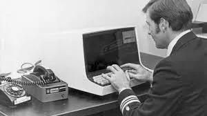 A black and white photograph of a man in a suit jacket with sleeve stripes typing on a vintage desktop computer terminal next to a rotary telephone.