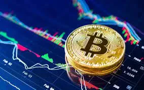 A physical golden Bitcoin token resting on a dark screen displaying multicolored cryptocurrency stock candle graphs and market data trends