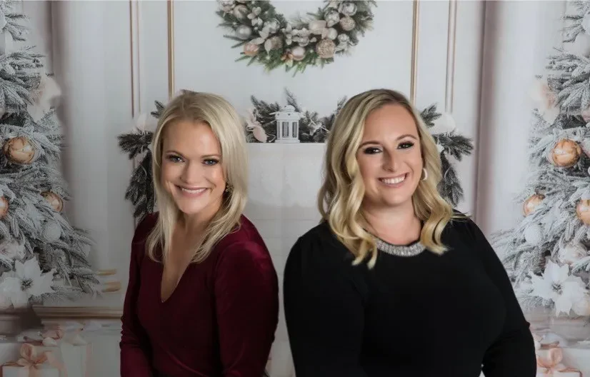  Attorneys Shari-Lynn Cuomo Shore and Kristen Wolf smile side-by-side in front of a festive holiday backdrop decorated with flocked Christmas trees and an elegant fireplace wreath