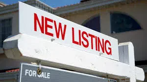 A white real estate sign with NEW LISTING printed in bold red letters, mounted above a smaller FOR SALE sign in front of a house.