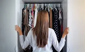 Rear view of a woman with long brown hair standing with her hands on the open doors of a white wardrobe closet filled with hanging clothes.