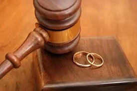 A wooden legal gavel resting on its sound block next to two gold wedding bands on a wooden tabletop.