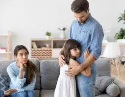 A young girl hugs her sad father tightly in a living room while her mother sits on the sofa in the background with a frustrated expression.