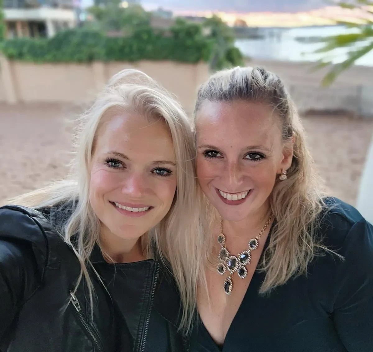 A close-up outdoor selfie of Shari Shore and Kristen Wolf smiling together at dusk with an open shoreline background.
