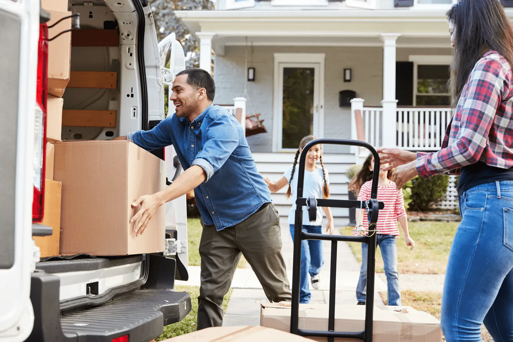 A family happily unloading cardboard moving boxes from the back of a white delivery van outside their new house, using a black hand truck.