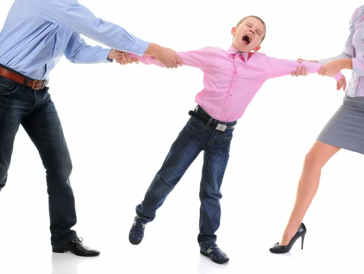A young boy in a pink shirt screams as two adults pull him in opposite directions by his arms against a plain white background.