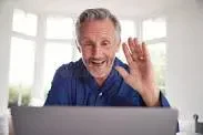 An older man with gray hair and a blue shirt smiles and waves at his laptop during a video call.