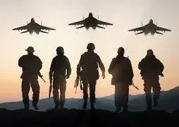 Silhouettes of five armed military soldiers walking forward in a line with three fighter jets flying overhead at sunset.