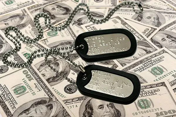 Two metal military identification dog tags with black rubber silencers resting on a scattered background of one-hundred-dollar bills