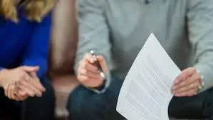 A close-up shot of a person holding a document and a pen while speaking to someone out of focus on the left.