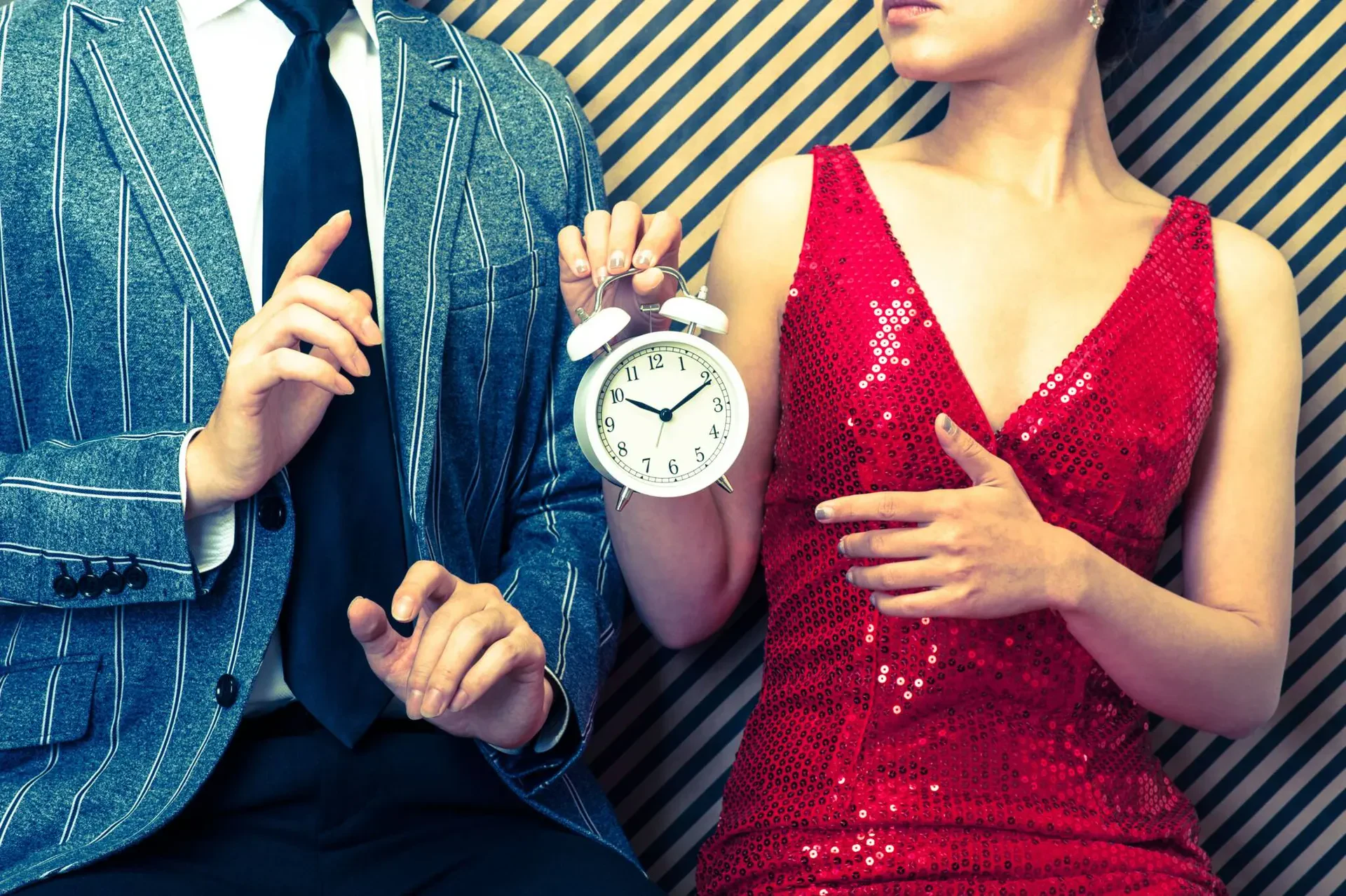 A man in a striped grey blazer and tie sits next to a woman in a sparkly red dress holding a white alarm clock.