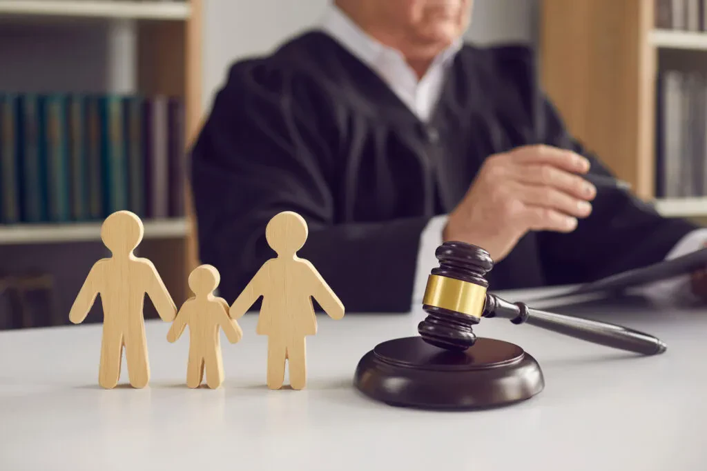 A legal gavel and wooden family cutouts on a desk with a judge in robes in the background.