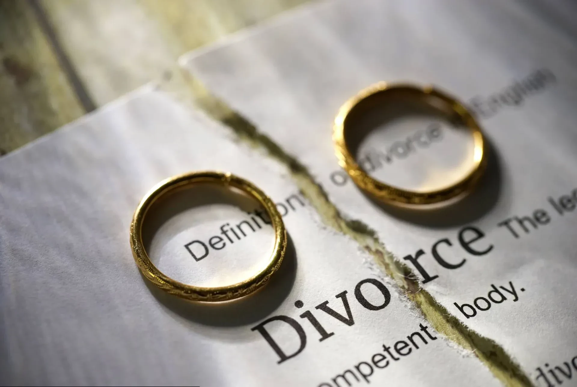 A close-up shot of a legal paper torn down the middle with the word "Divorce" printed on it, featuring two gold wedding rings placed on either side of the tear.