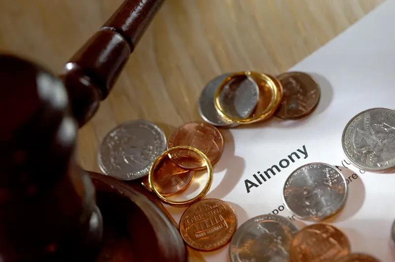 A gavel, two gold rings, and assorted coins rest on a document labeled "Alimony," symbolizing legal and financial aspects of divorce.